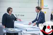 Kaksi henkilöä keskustelemassa teollisessa ympäristössä kuljetinhihnan äärellä. Taustalla näkyy IT-Line-logo ja Manutecin maskottihahmo Manu.